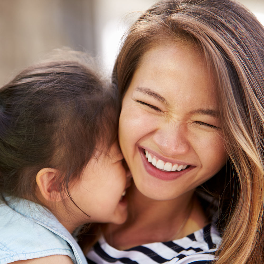 An adult woman with a bright smile is hugging a young child who is also smiling and looking up at her.